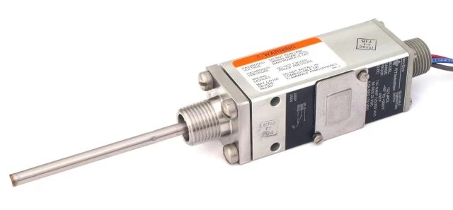 Close-up of a compact temperature switch. The switch is silver, with a probe extending from one end. It has a warning label and electrical connections.