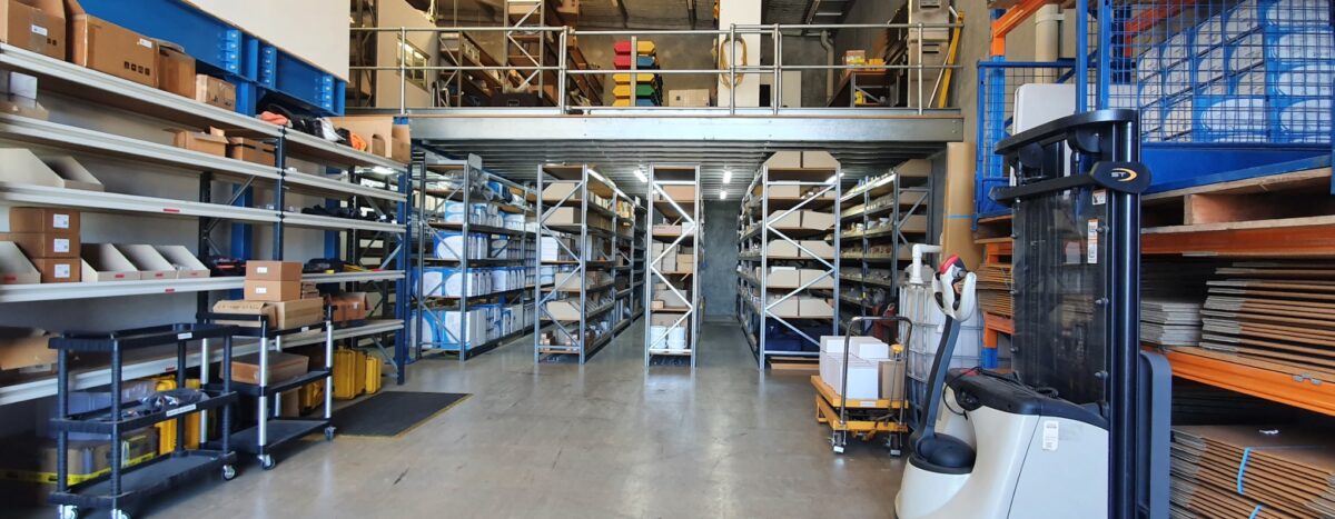 About us: Warehouse interior with shelving, boxes, and a forklift. Mezzanine level visible. Storage and distribution center.